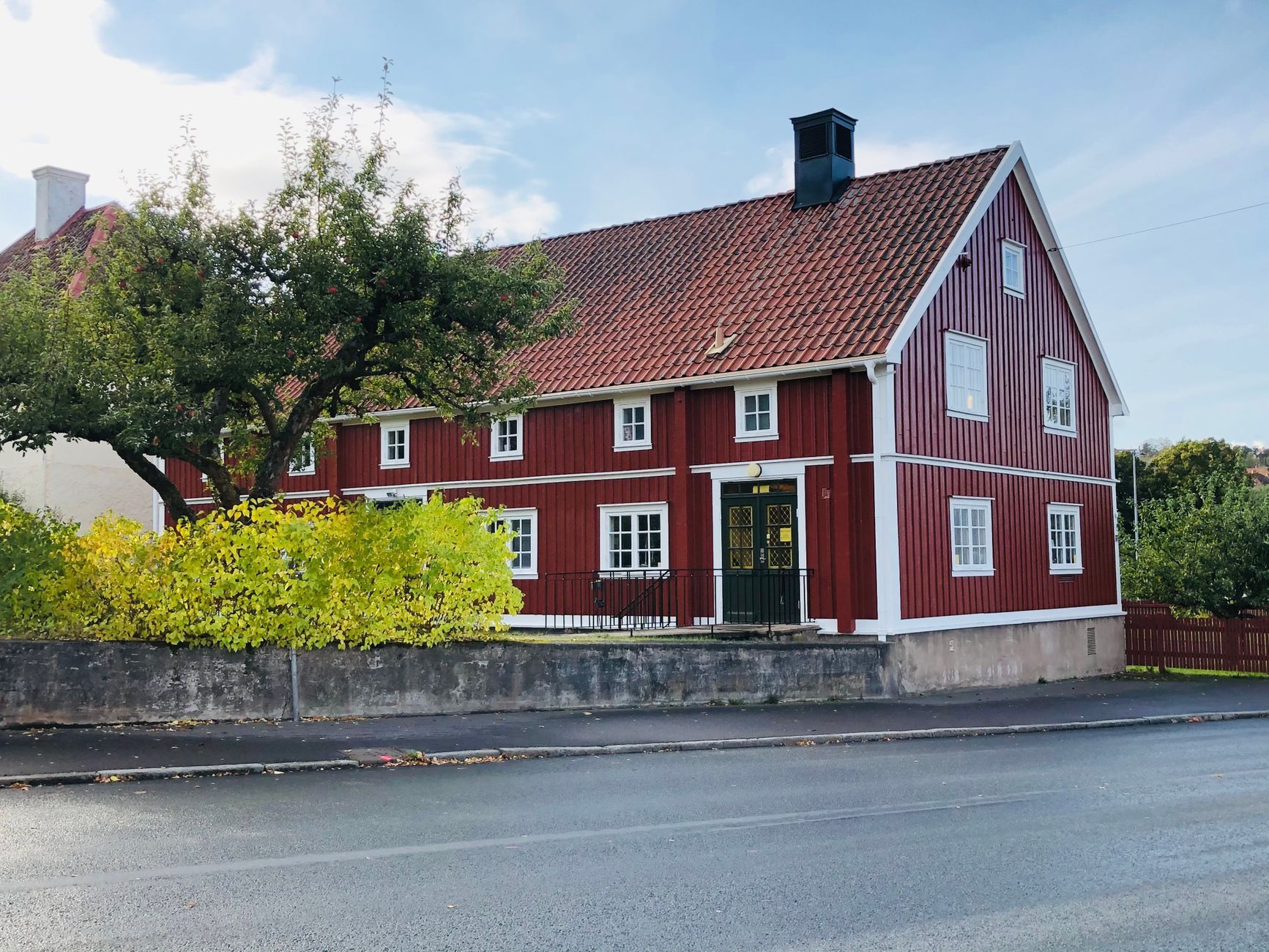 Rødt trehus med hvite vinduskarmer bak en lav mur og et tre