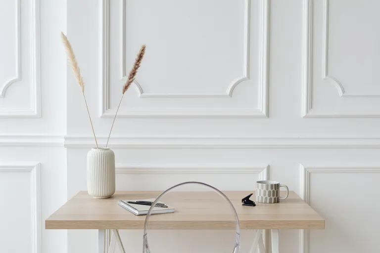 Lyst skrivebord i tre med notatbok, penn, kopp og en vase med tørkede strå
