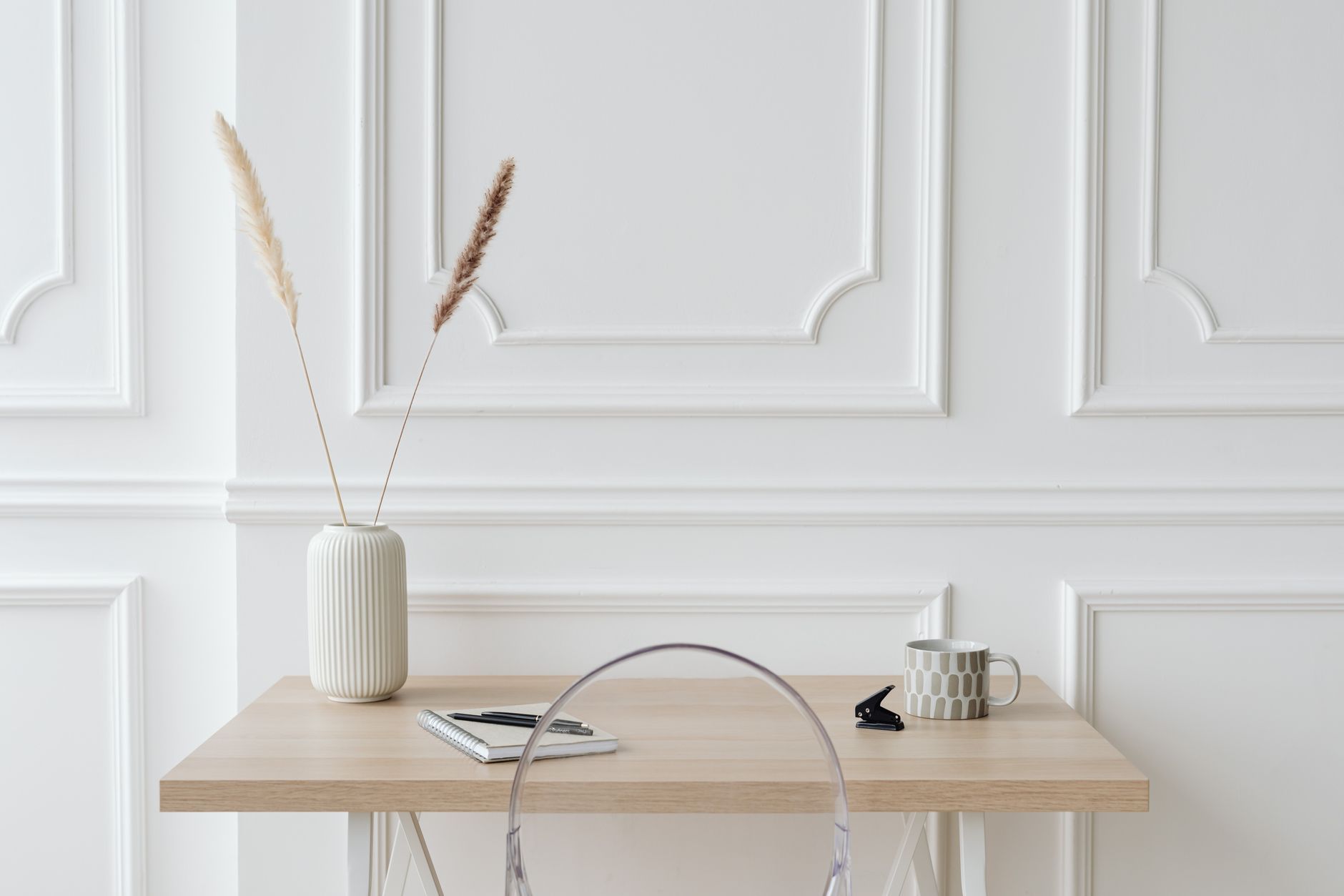 Lyst skrivebord i tre med notatbok, penn, kopp og en vase med tørkede strå