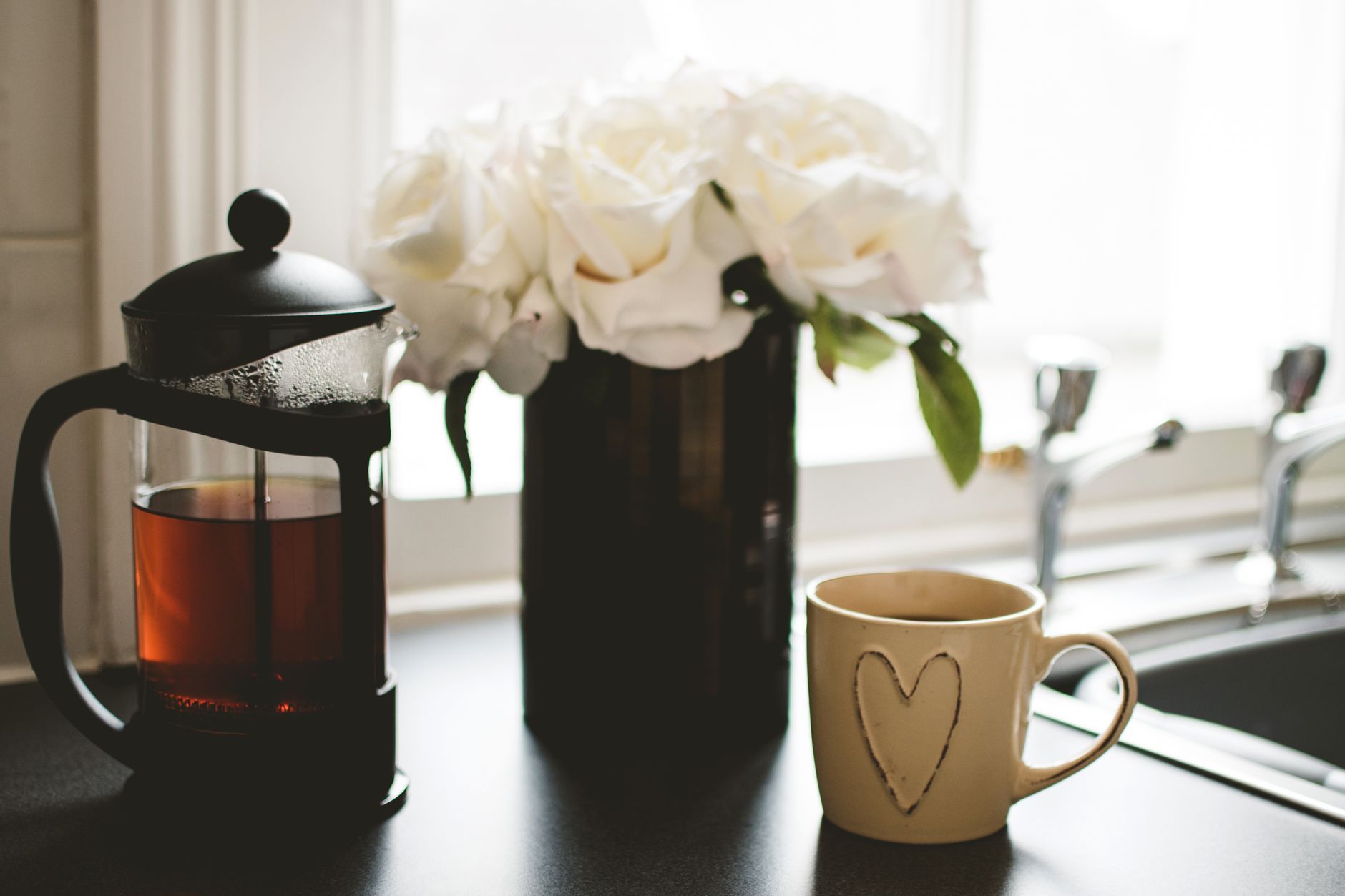 Kaffekopp, presskanne og hvite roser i et kjøkkenvindu i morgenlys