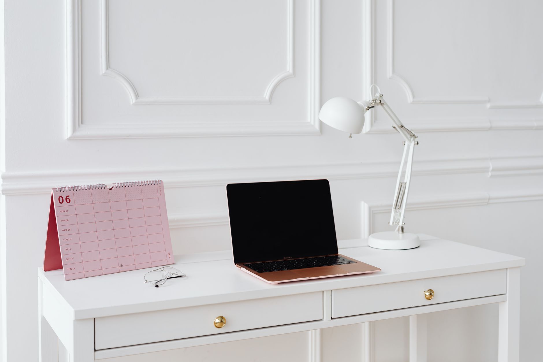 Hvitt skrivebord med bordkalender, lukket laptop og skrivebordslampe
