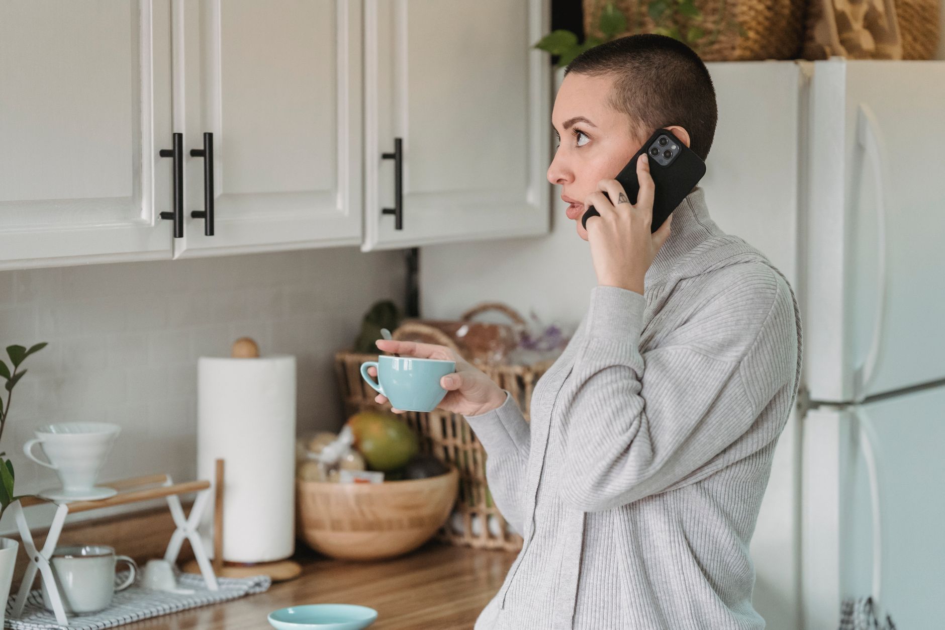 Kvinne i grå genser snakker i telefonen med en kaffekopp på kjøkkenet